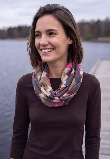 Loop-sjaal met grafische print truffle brown Jonge vrouw lacht op een steiger bij het water, draagt een donker shirt en een kleurrijke sjaal.