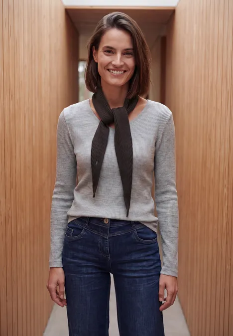 Eine lächelnde Frau in grauem Pullover und Jeans vor Holzpaneelen.