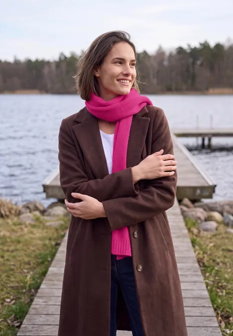 Lange gebreide sjaal in effen kleur pink flash Een lachende vrouw in een bruine jas en roze sjaal staat op een steiger bij een meer.
