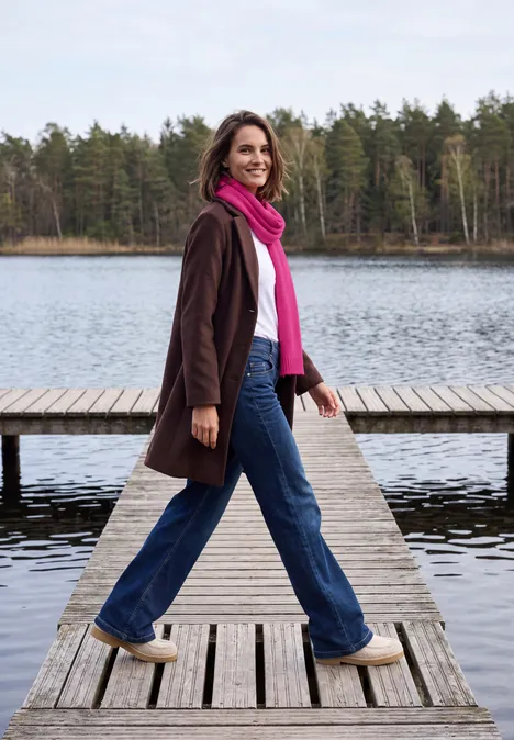 Lange gebreide sjaal in effen kleur pink flash Vrouw met jas en sjaal wandelt glimlachend over een houten steiger langs een meer met bos op achtergrond.