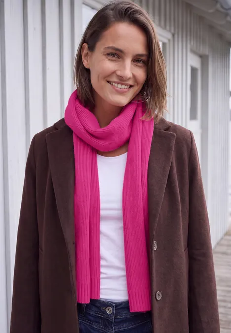 Lange gebreide sjaal in effen kleur pink flash Een glimlachende vrouw in een bruine jas en roze sjaal voor een wit gebouw.