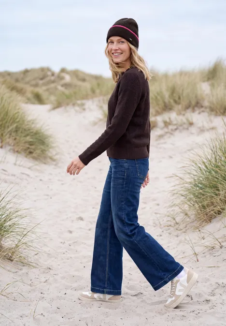 Femme en pull noir, jean large, bonnet et baskets blanches se promène sur une plage de dunes.