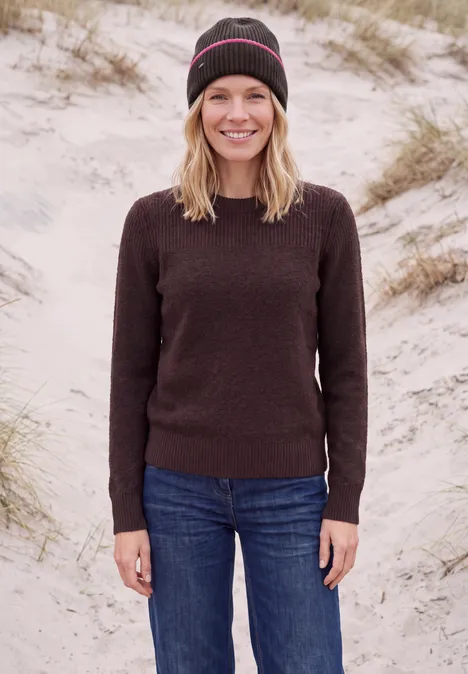 Une femme en bonnet, pull et jean sourit sur une plage de dunes.