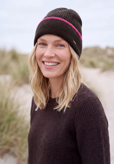 Une femme souriante coiffée d'un bonnet sur la plage devant des dunes.