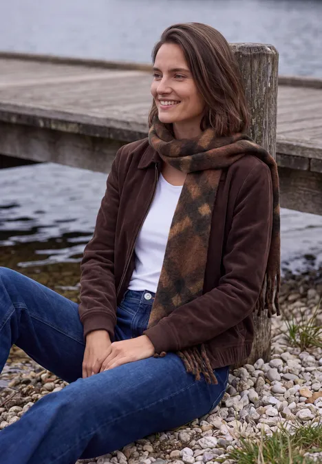 Een geruwde sjaal met franjes mocha brown Frau in Jeans, brauner Jacke und Schal sitzt lächelnd am Ufer auf Kies.