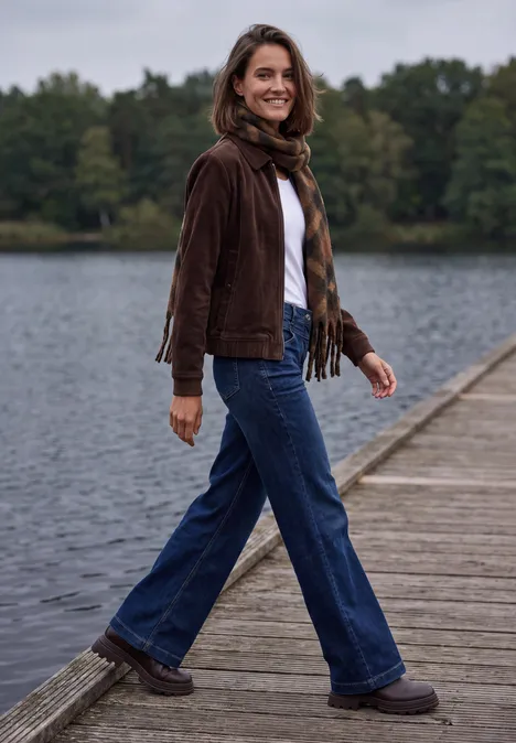Een geruwde sjaal met franjes mocha brown Eine Frau in brauner Jacke und Jeans geht lächelnd auf einem Holzsteg am See entlang.