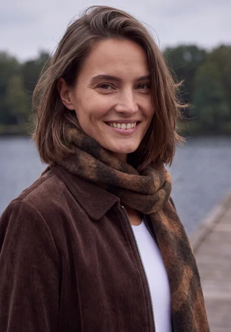 Een geruwde sjaal met franjes mocha brown Eine lächelnde Frau in braunem Mantel und Schal an einem Seeufer.