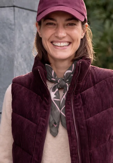 Une femme souriante porte une veste bordeaux, un foulard et une casquette devant un mur en pierre.