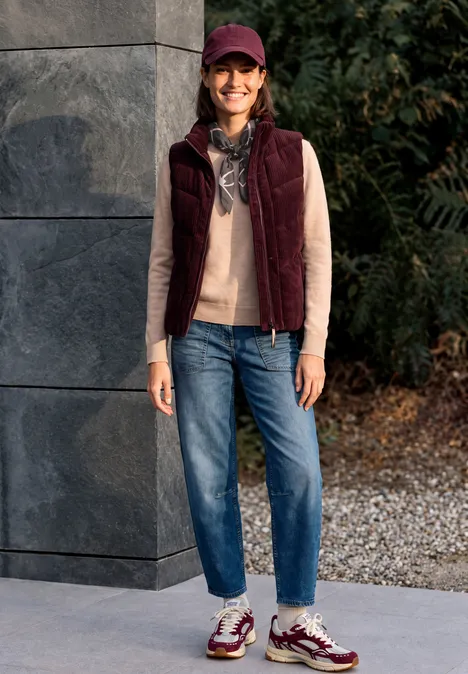 Femme souriante en casquette, écharpe, pull beige, veste bordeaux et jean, debout devant un mur gris.