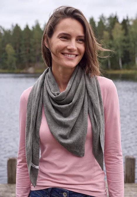 Lächelnde Frau in rosa Pullover und grauem Dreieckstuch am See im Wald.