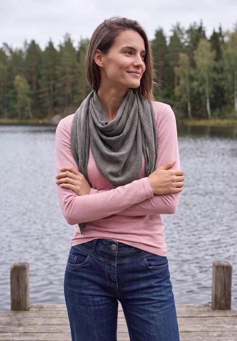 Eine lächelnde Frau mit rosa Pullover und Schal steht am Seeufer vor einem Wald.