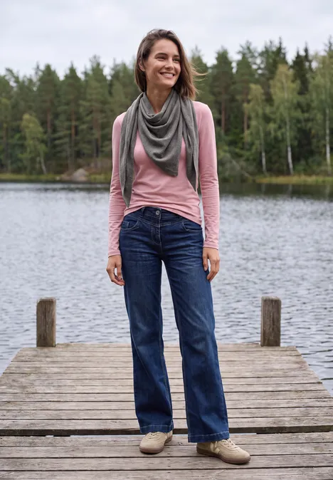 Frau steht lächelnd auf einem Steg am See, trägt Jeans, rosafarbenes Shirt und Schal, bewaldeter Hintergrund.