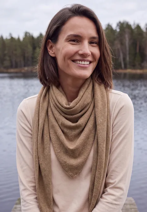 Lächelnde Frau mit braunem Schal sitzt am See, Wald im Hintergrund.