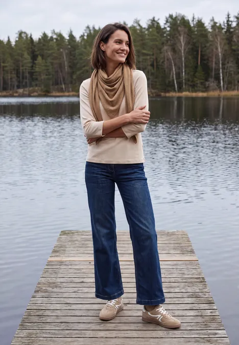 Frau mit Schal und Jeans auf Steg am See, umgeben von Wald, lächelt und schaut zur Seite.
