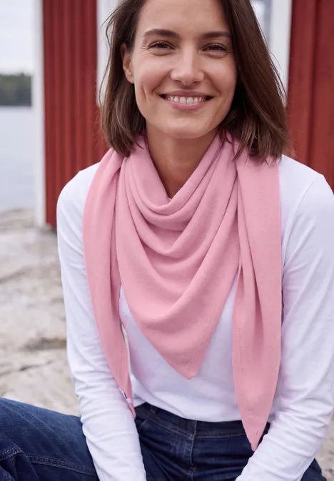 Eine lächelnde Frau in weißem Shirt und rosa Dreieckstuch sitzt am Wasser.