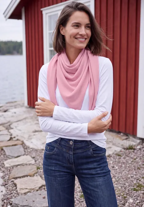 Eine Frau mit rosa Schal und weißem Oberteil steht lächelnd vor einem roten Holzhaus am See.