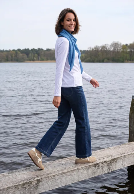 Dreieckstuch in Unifarbe north blue melange Frau mit blauem Schal, Jeans und Sneakern spaziert auf einem Steg am See und lächelt zurück.