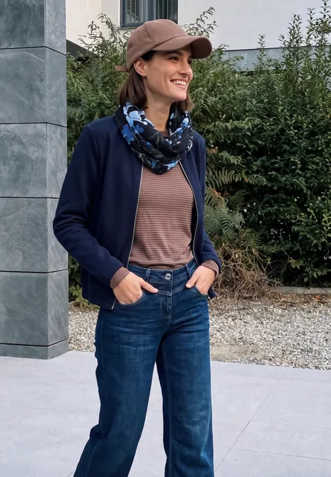 Eine lächelnde Frau in Jeans, Cardigan, Tuch und Baseballcap steht draußen vor einem Gebäude.