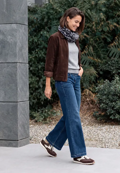 Frau in brauner Jacke, Jeans und Halstuch läuft an einem modernen Gebäude mit Säule und Grünpflanzen vorbei.