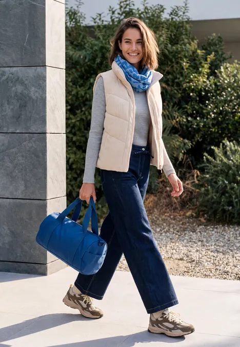 Écharpe Loop avec motif dévoré north blue Frau in Freizeitkleidung mit einer blauen Reisetasche, lächelnd vor einem modernen Haus.