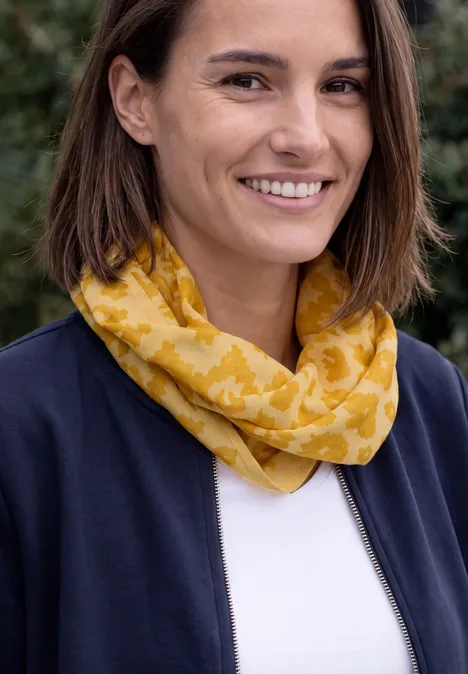 Écharpe Loop avec motif dévoré golden honey Lächelnde Frau mit gelbem Schal und Jacke vor unscharfem, grünen Hintergrund.
