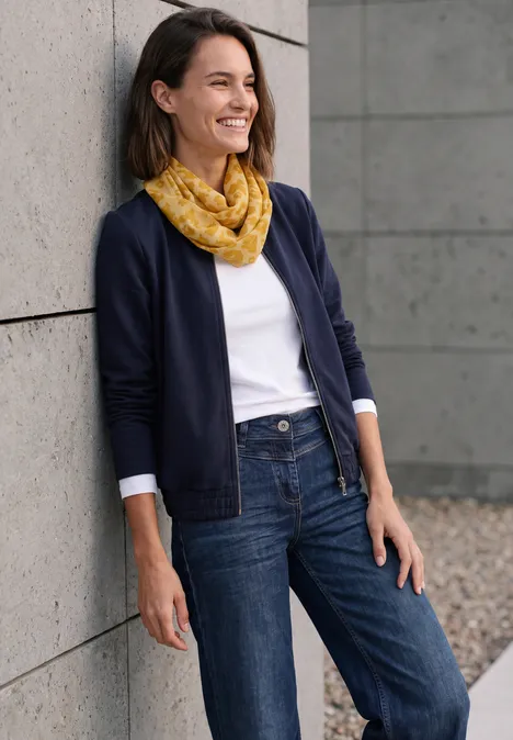 Écharpe Loop avec motif dévoré golden honey Eine lächelnde Frau lehnt im Freien an einer Wand, trägt Jeans, weißes Shirt, dunkelblauen Cardigan und gelben Schal.