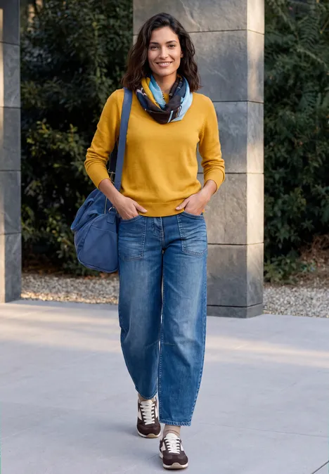 Écharpe Loop à imprimé graphique truffle brown Une femme souriante porte un pull jaune, un jean bleu, une écharpe et un sac bleu en marchant en plein air.