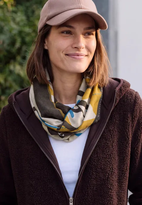 Frau mit brauner Jacke, Cap und gemustertem Schal, lächelt leicht im Freien.