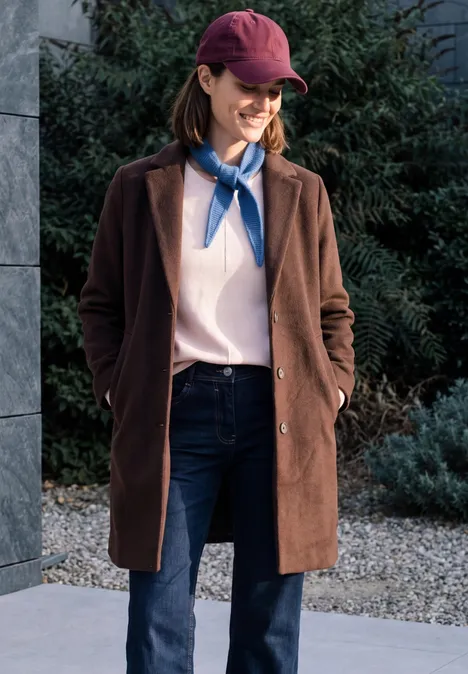 Frau im roten Cap, braunem Mantel, Jeans und blauem Halstuch, steht draußen und lächelt.
