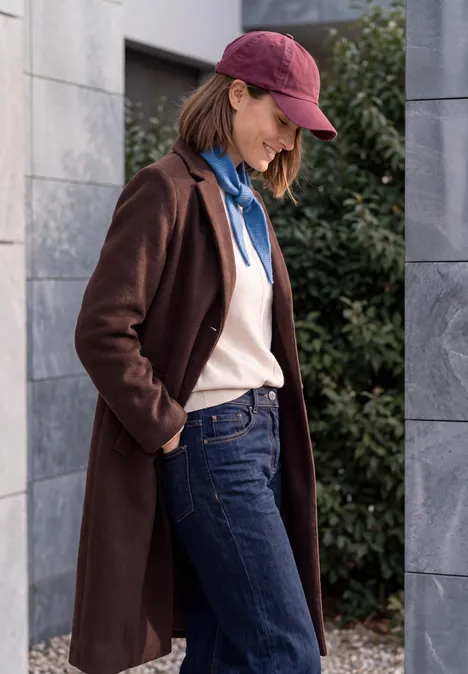 Frau in Jeans, Pullover, blauem Hoodie, braunem Mantel und roter Cap steht draußen vor einer grauen Steinwand.