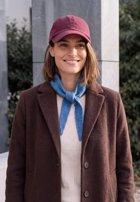 Lächelnde Frau mit rotem Cap, braunem Mantel und blauem Halstuch vor urbanem Hintergrund.