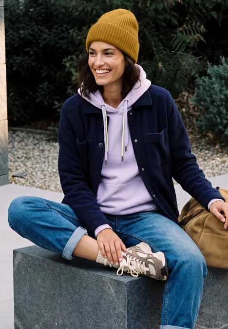 Effen muts golden honey melange Junge Frau in Mütze, Hoodie, Jeans und Sneakers sitzt lächelnd im Freien auf einer Mauer.