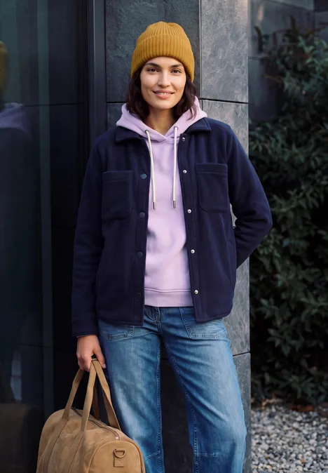Effen muts golden honey melange Eine junge Frau steht draußen vor einem Gebäude, trägt eine gelbe Mütze, rosa Hoodie, Jeans und hält eine Tasche.