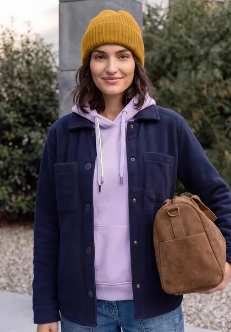 Effen muts golden honey melange Junge Frau mit gelber Mütze und lila Kapuzenpullover trägt eine braune Tasche vor Grünpflanzen.