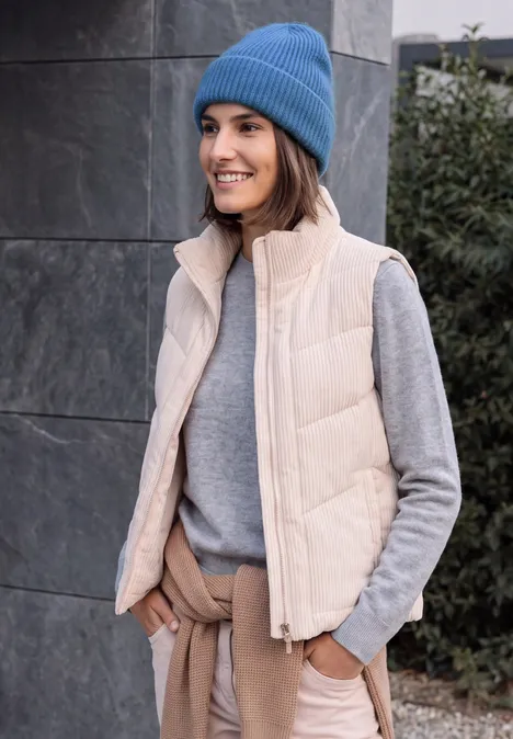 Frau in blauer Mütze, grauem Pullover und beiger Weste lächelt mit den Händen in den Taschen.