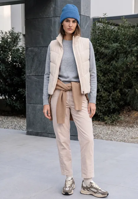 Frau in lässiger Outdoor-Kombi: blaue Mütze, grauer Pullover, beige Weste und Hose, Sneaker.