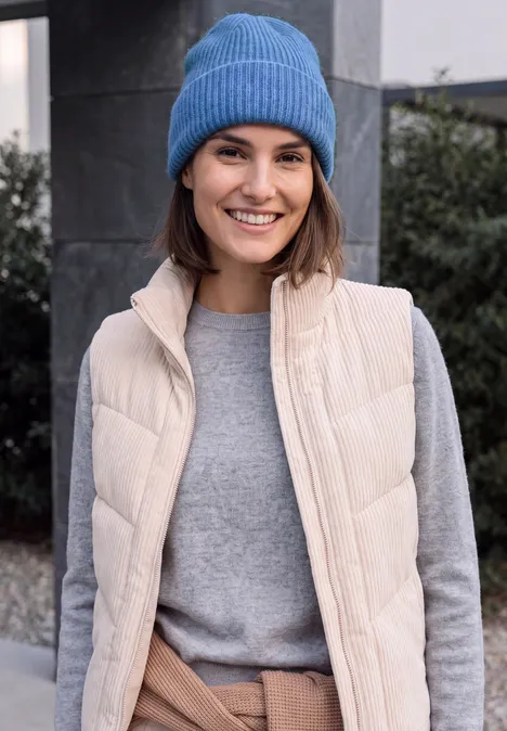 Eine lächelnde Frau mit blauer Mütze, beiger Steppweste und grauem Pullover im Freien.