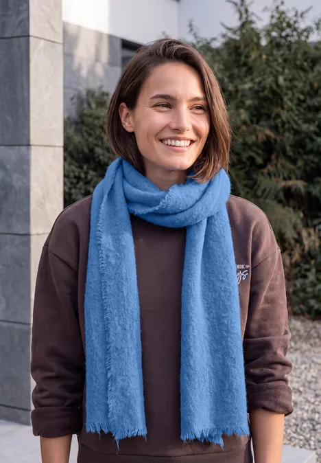Kuscheliger Schal in Unifarbe north blue Junge Frau mit braunem Pullover und blauem Schal lächelt im Freien vor modernem Gebäude.