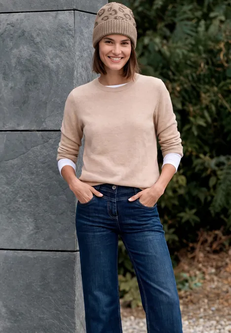 Une femme souriante en pull et bonnet beiges, jean bleu, posant devant un mur gris et des buissons verts.