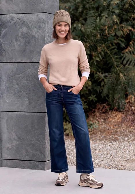 Jeune femme en bonnet et pull beige, jean ample et baskets, posant devant un mur de pierre et un décor végétal.
