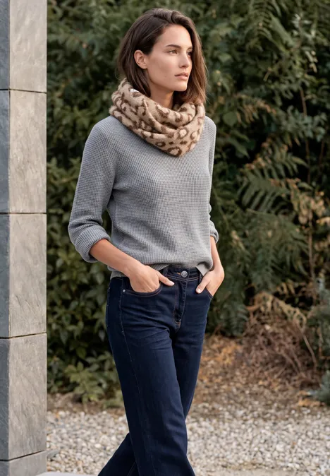 Femme en pull gris et écharpe, jean foncé, mains dans les poches, debout près d'une colonne.