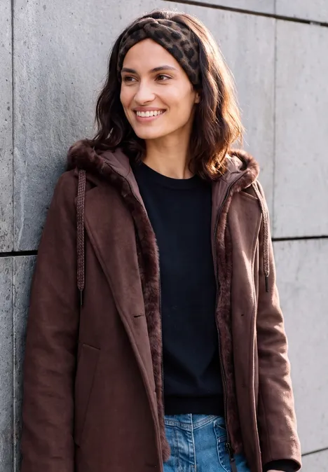 Eine lächelnde Frau mit braunem Mantel, schwarzem Shirt und Jeans lehnt an einer grauen Wand.