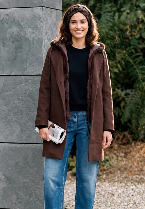 Eine lächelnde Frau in braunem Mantel, schwarzem Pullover und Jeans steht draußen und hält ein Fernglas.