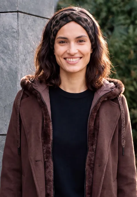 Eine lächelnde Frau mit brauner Jacke, schwarzem Shirt und Haarreif vor einer Steinmauer.