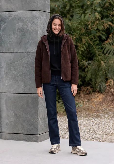 Balaclava mit Leo-Muster truffle brown Frau in brauner Jacke, Jeans und Sneakern lehnt lächelnd an grauer Wand im Freien, umgeben von Grün.