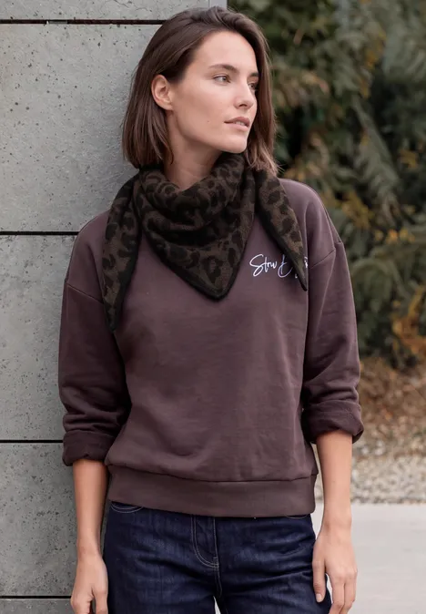 Femme appuyée sur un mur en béton, portant un pull marron foncé et un foulard à motif.