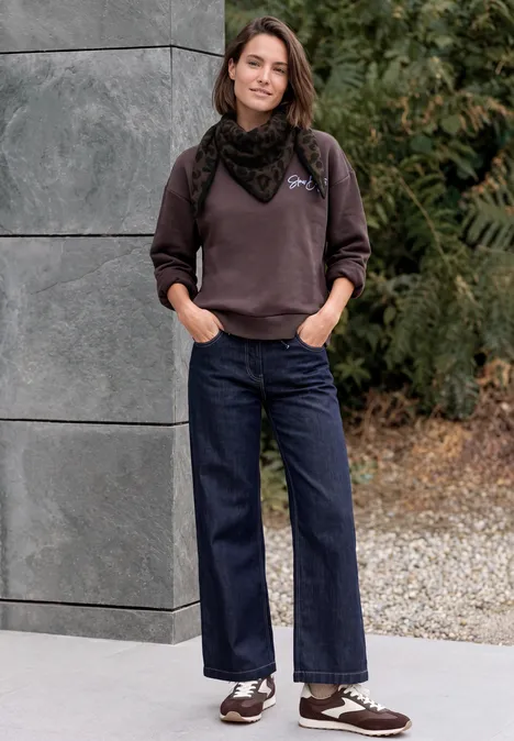 Une femme en pull, foulard et jeans larges, mains dans les poches, pose décontractée devant un mur et des plantes.