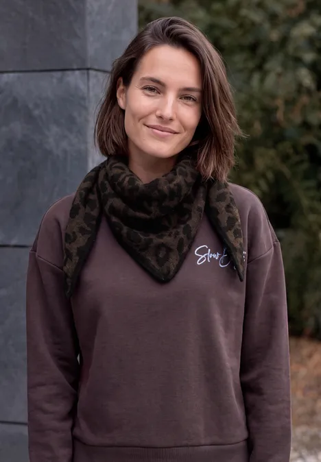 Jeune femme souriant, pull marron et écharpe à motif, en extérieur devant un mur en pierre et des plantes.