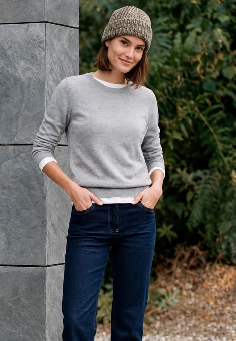 Junge Frau mit grauem Pullover, Jeans und Mütze lehnt lässig an einer Mauer im grünen Außenbereich.