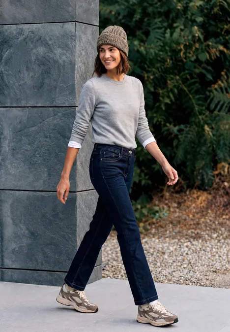 Frau in Jeans, Pullover und Mütze läuft entspannt an einer Steinwand und grünen Hecken vorbei.